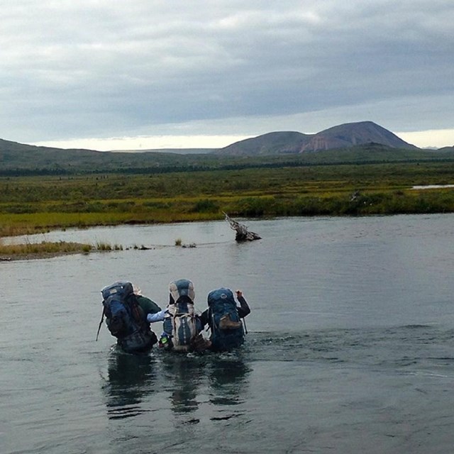 3 backpackers wade through a river