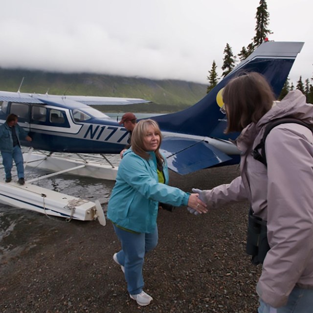 2 women shaking hands with a two men standingnear a float plane in the background.