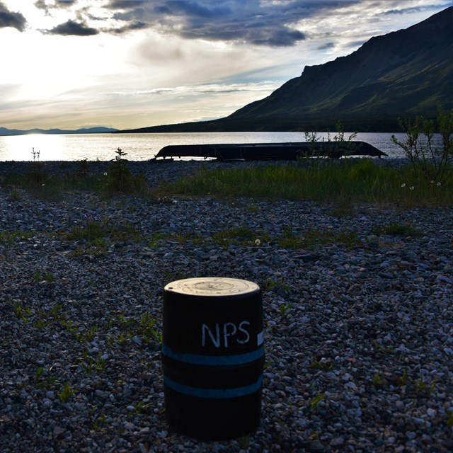 a bear barrel sits on a beach with distant kayak and mountains at dusk in the background