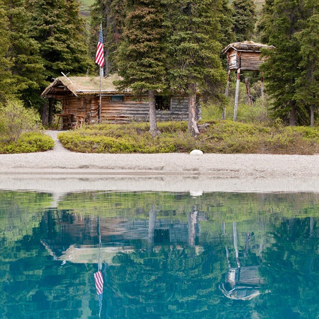 Photograph of a small log cabin nestled in the woods reflecting in a blue-green lake.