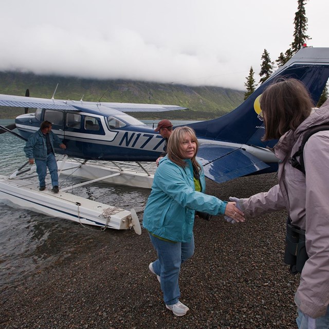 two women shake hands on a beach with a floatplane in the background