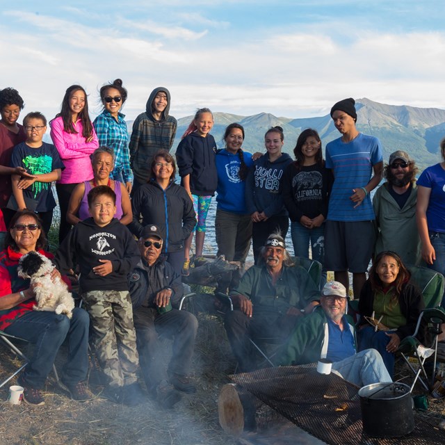 A group of people of all ages pose in front of a camp fire with water and mountains behind them.