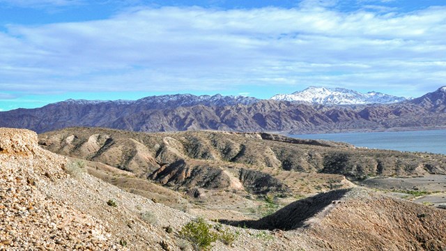 A person stands on the edge of a hilly overlook. Water and mountainous landscape in background.