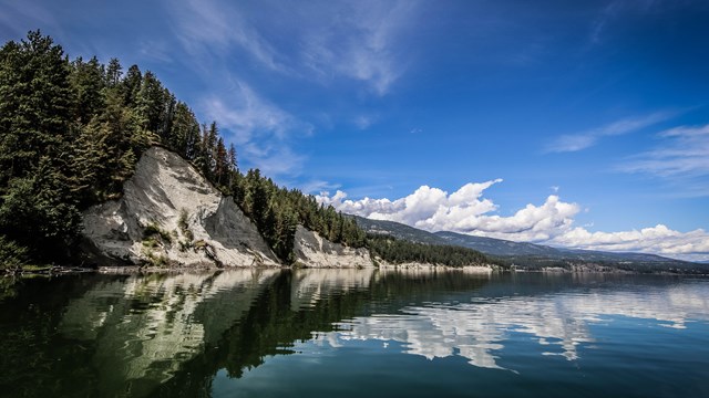 A view down the lake with calm water reflecting white cliffs, pines, clouds and sky.