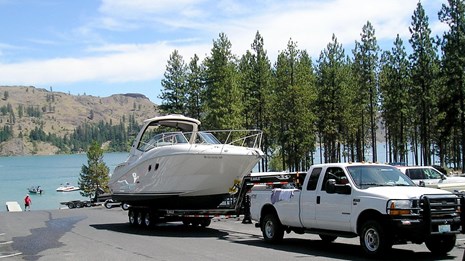 a white truck backs a large white boat on a trailer down a boat launch ramp.