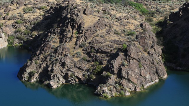 Aerial view of sloping cliffside of brown rocks emerging out of bright blue water.