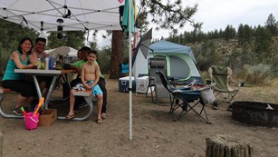 A family enjoying a camping trip, sitting at a picnic table under a shade shelter.