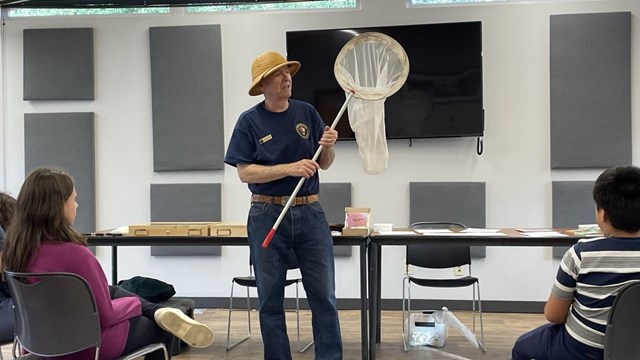 A man in a blue shirt and a tan hat holds a bug net as he speaks to an audience.