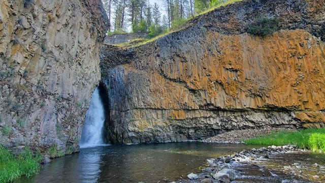 An arcing waterfall squeezes itself out of a crack between brown, orange, and grey rock, into a pool