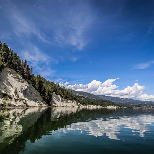 A view down the lake with calm water reflecting white cliffs, pines, clouds and sky.