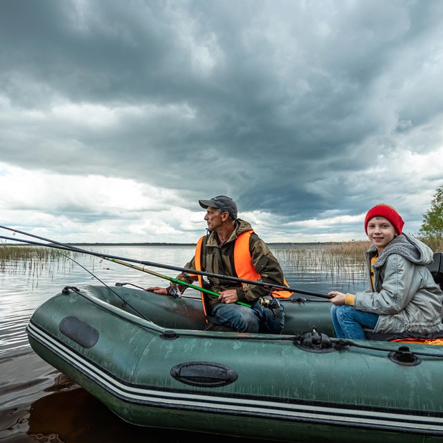 Two people fishing along the shoreline in an inflatable boat.