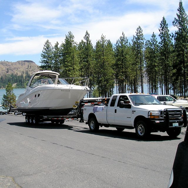 a white truck backs a large white boat on a trailer down a boat launch ramp.