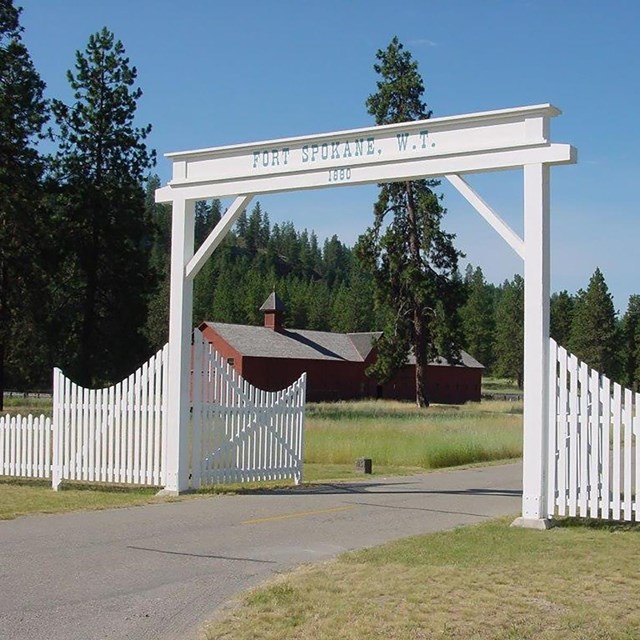 The historic wooden gateway at Fort Spokane with the large red mule barn beyond.