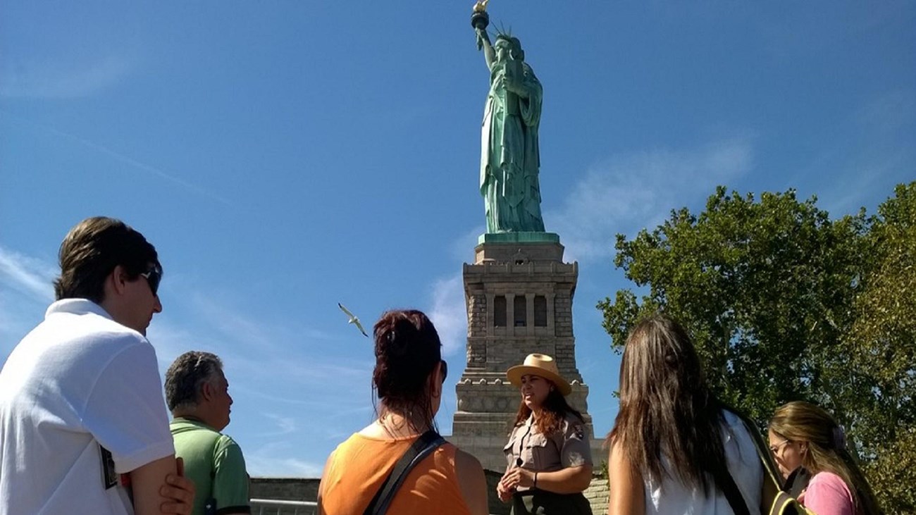 Several visitors listen as a park ranger talks about the history of the site, with the Statue of Lib