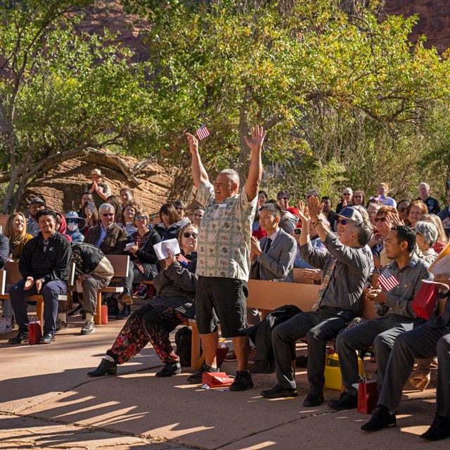 A man in dark shorts and a patterned shirt stands with his arms raised overhead among a crowd 