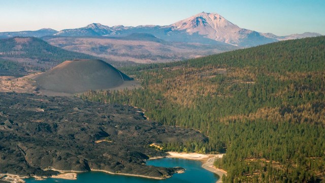 An aerial photo of a volcanic landscape including a lava flow, volcanic peaks, and conifer forests s