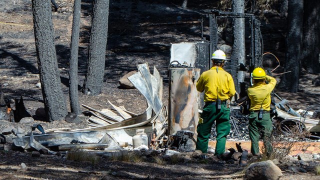 Two people in yellow shirts, green pants, and hard hats record data of a destroyed structure backed 