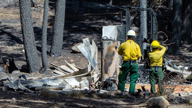 Two people in yellow shirts, green pants, and hard hats record data of a destroyed structure backed 