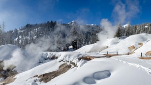 A snow-covered road passes through steaming hydrothermal features in a mountain landscape.