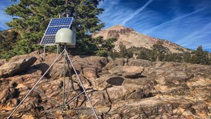 A rounded device sits atop a tripod on rocky ground, backed by a solar panel and mountain