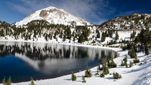 A panoramic photo of a snow-covered peaks reflected in a blue lake lined by a thin layer of snow.