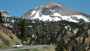 A white car on a winding highway surrounded by steep slopes and a large, grey mountain.