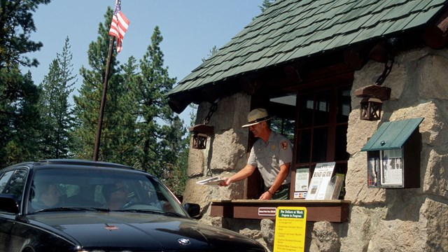 A man in a park ranger uniform hands a person in a vehicle from inside a small stone building