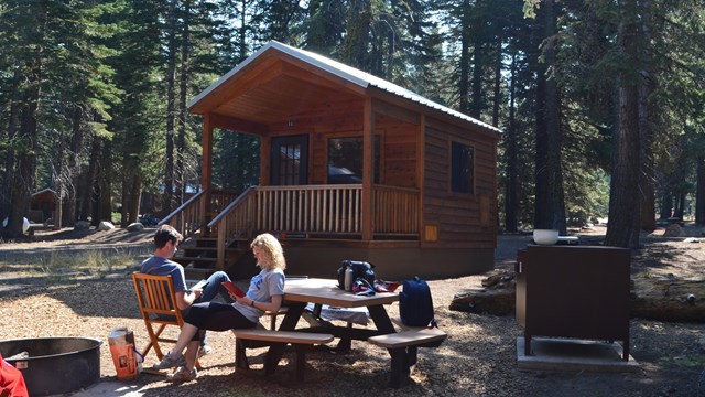 A man a woman sit and read outside of a small, wooden cabin
