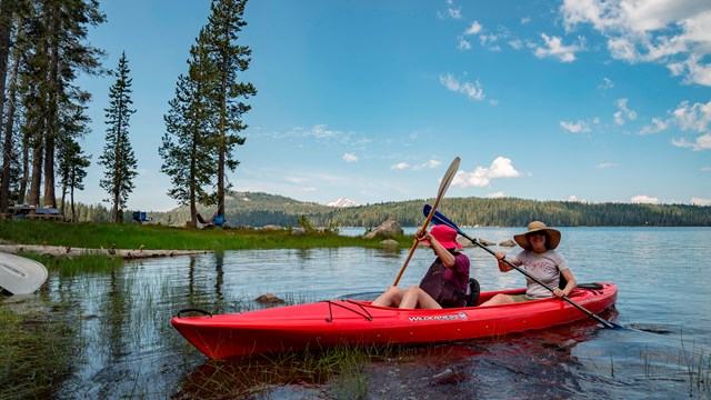 Two women sit in a red kayak on a lake backed by a snow-capped mountain