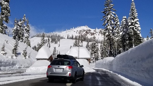 A silver wagon drives toward two small, snow-covered buildings.