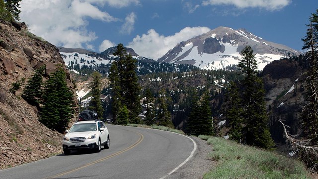 A white vehicle on a highway backed by a snow-covered volcano