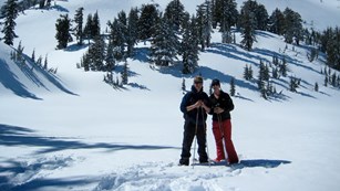 A man and a women standing on snowshoes backed by a pointed, snow-covered peak.