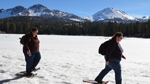 Two women on snowshoes walk across the shore of a snow-covered lake backed by volcanic peaks.
