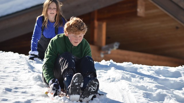 A boy slides down a hill backed by a young girl and the top of a wooden building.