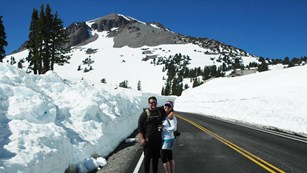 A man and woman stand on a road flanked by snow