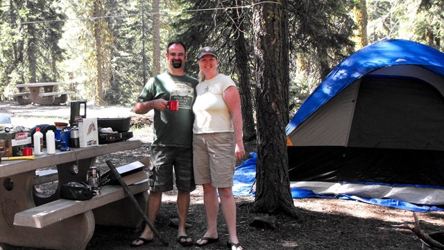 A man and a woman standing next to a picnic table and tent at a campground.