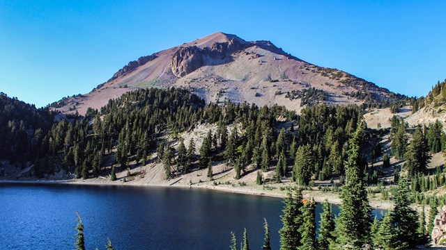 A lake sits at the base of a large mountain covered in pine trees on a clear sunny day. 