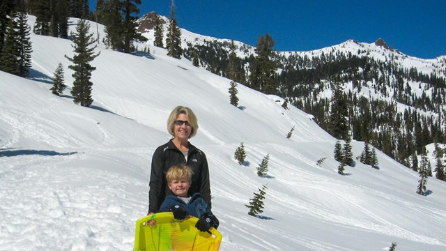 A woman and young boy stand with a yellow sled in a snowy mountain landscape.