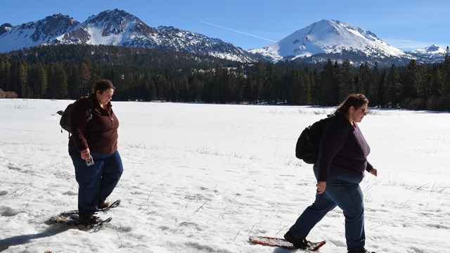Two women on snowshoes walk across the shore of a snow-covered lake backed by volcanic peaks.