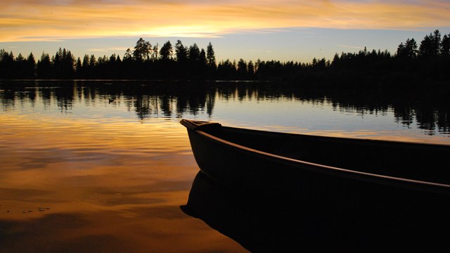 Half of a canoe in a lake lined by conifer trees under an orange sunset sky