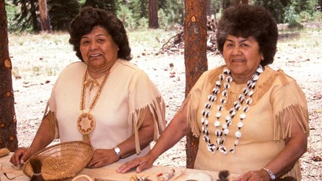 Two women in Native American attire stand at a table of traditional Indian items.