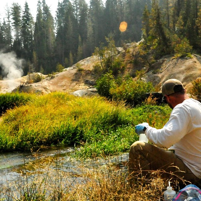 A man holds a collection bottle on the edge of a creek with stream rising in the background.