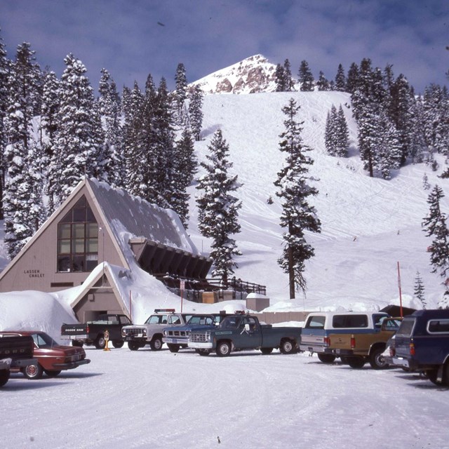 Vehicles parked in a snow-covered parking area next to a two-story a-frame chalet at the base of a s