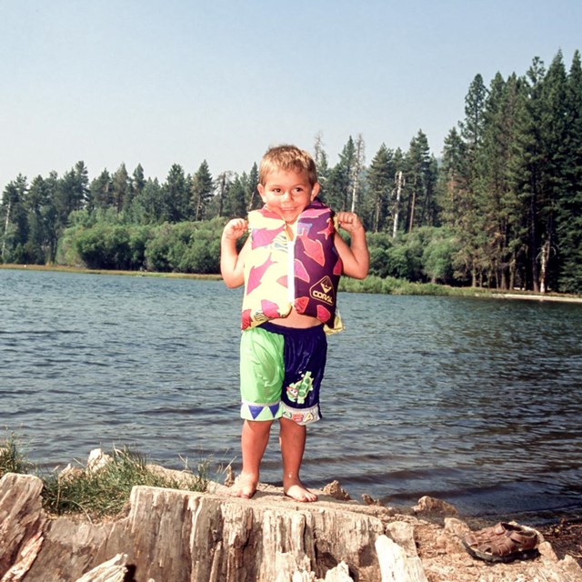 A boy in a life vest stands poses for a photo on a tree stump in front of a lake.