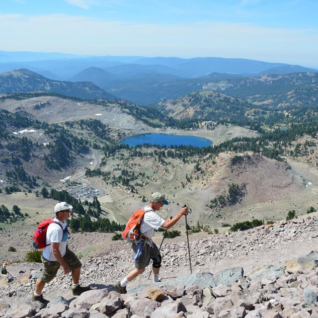 Two men hike on a rocky trail high above a parking are and blue lake surrounding by mountains.