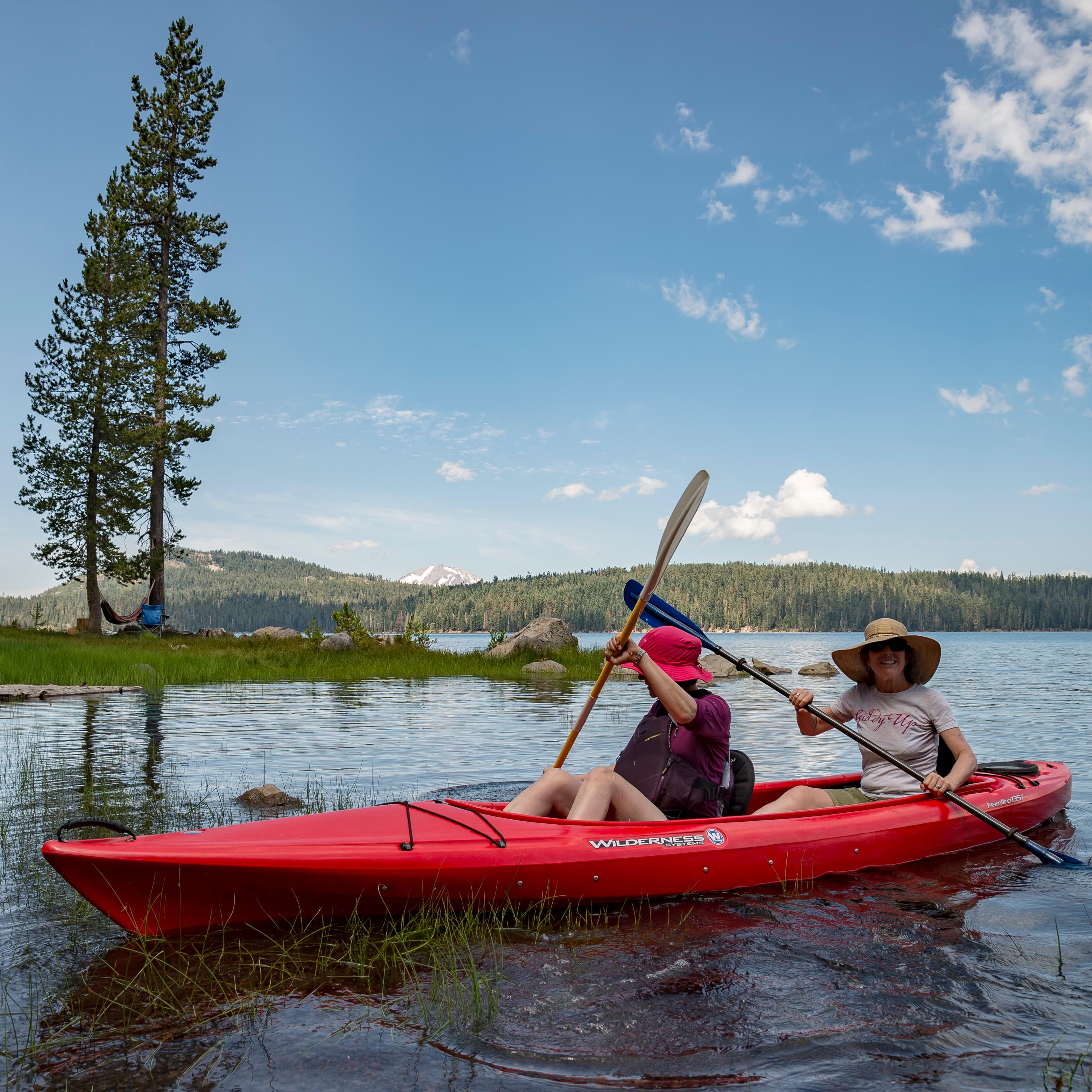 Two women sit in a red kayak on a lake backed by a snow-capped mountain