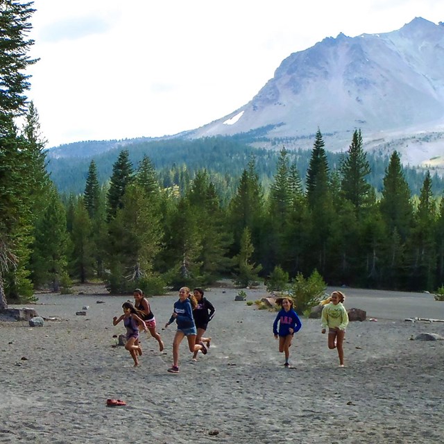Eight girls run across a gray sandy area at the base of a volcano.