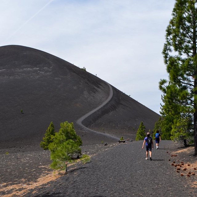 Two hikers on a sandy trail at the bottom of a barren, cone-shaped volcano.