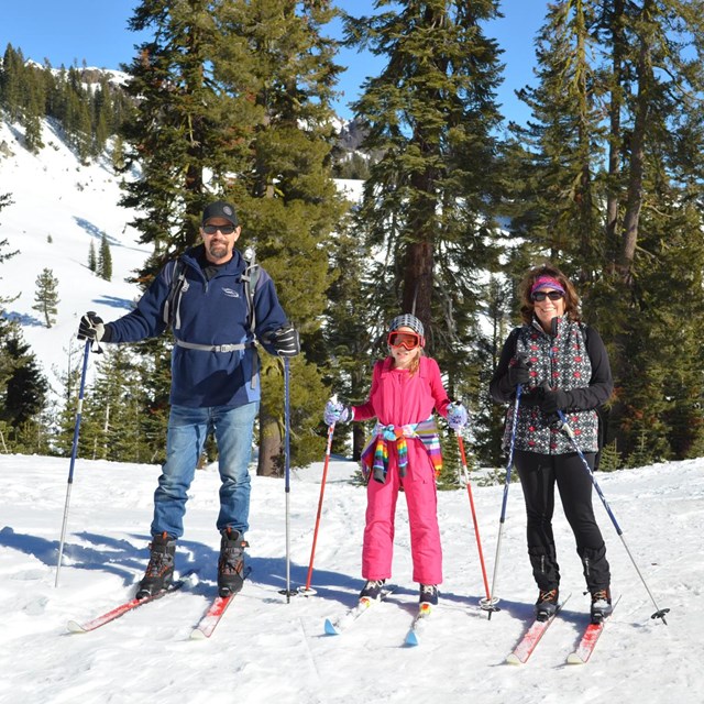 A man, woman, and young girl on cross-country skis in a mountain landscape.