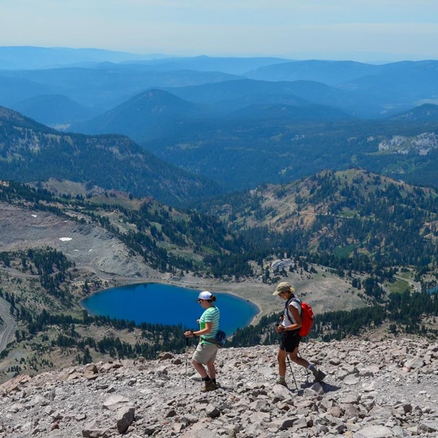 Two people hike on a rocky trail above a mountain landscape including a blue lake and numerous volca